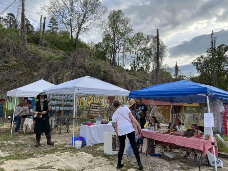On a hillside lot overgrown with vines and weeds, a flat place has been cleared and three artists' booths are set up with their creative works for sale. Customers and one person dressed in roguish garb are visiting the booths.